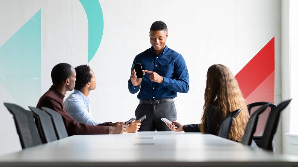 Young professional presenting mobile device to diverse team during business meeting at modern office.