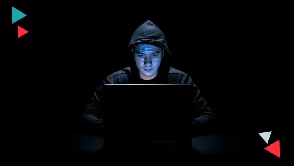 Young hacker in hoodie working on a laptop in a dark setting, representing cybersecurity threats.