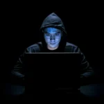 Young hacker in hoodie working on a laptop in a dark setting, representing cybersecurity threats.
