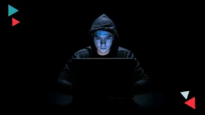 Young hacker in hoodie working on a laptop in a dark setting, representing cybersecurity threats.