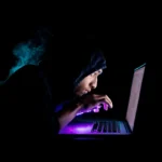 Young Hacker working on a laptop in a dark setting, emphasizing cybersecurity and IT security services.