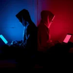 Two hackers in hoods sit back-to-back, each using a laptop in blue and red neon lighting, creating a cyber-themed scene.