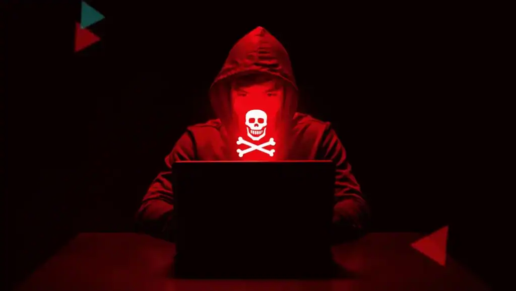 Hacker in red hoodie with skull mask working on a laptop in a dark environment, representing cybersecurity threats.