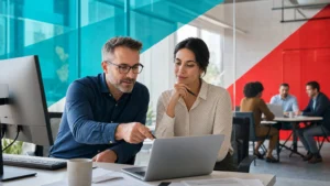 Two business colleagues reviewing shared company documents on a laptop and monitor in a bright modern office