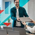 IT services professional handing over a laptop in a modern office setting.