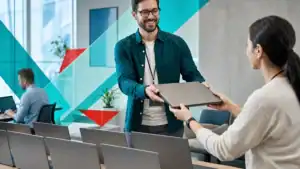 IT services professional handing over a laptop in a modern office setting.
